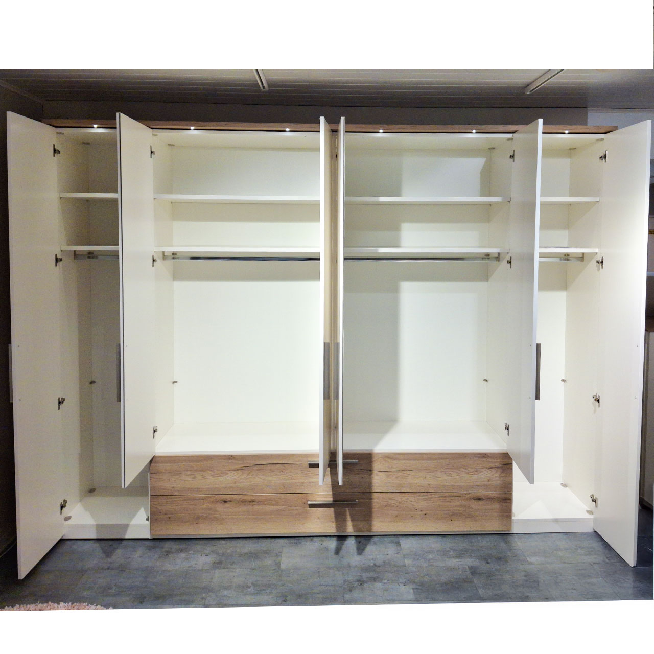 Ein 6-türiger Kleiderschrank mit weißen Fronten und Griffen in Edelstahl. Die Türen sind alle geöffnet. Im unteren Bereich sind zwei Schubkästen, die über die Breite von vier Schranktüren reichen. Die Fronten der Schubkästen sind in Eiche.