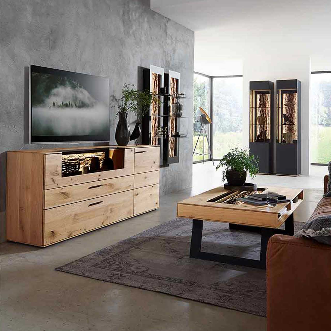 Hartmann Runa - Sideboard 8410-4191 - Milieu Wohnzimmer, in dem rechts ein Sideboard aus hellem Holz steht und rechts auf einem Teppich ein Couchtisch auf Kufen vor einem Sofa. Im Hintergrund sind zwei Hängeelemente und zwei Standvitrinen zu sehen.