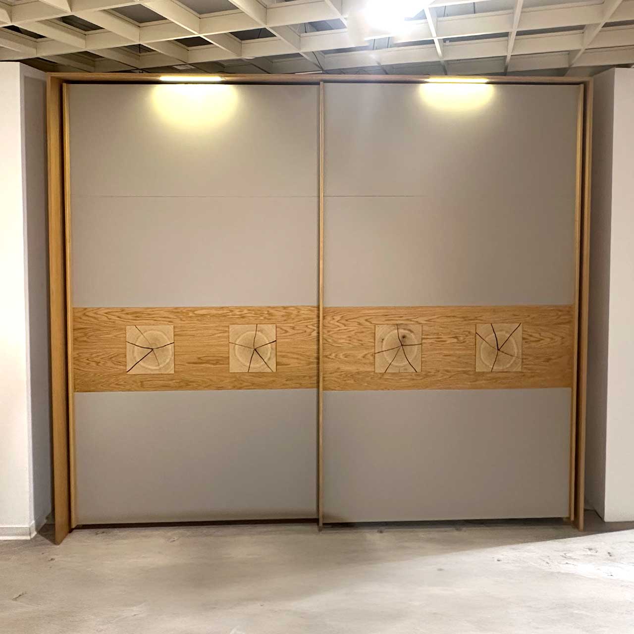Der Außenkorpus ist aus Holz, die zwei grauen Türen tragen als Zierde ein Holzband, auf dem pro Tür zwei quadratische Hirnholzapplikationen eingearbeitet sind. Oben am Rahmen findet sich je Tür eine Lichtquelle, die den Schrank beleuchtet.