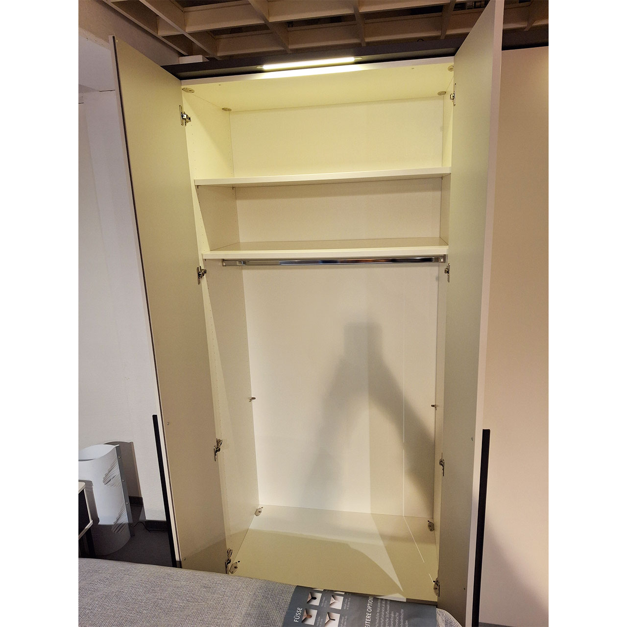 Mehrtüriger Kleiderschrank mit LED-Beleuchtung über jeder Doppeltür. Die Doppeltür ganz links ist geöffnet. Zu erkennen sind eine Kleiderstange und zwei Einlegeböden im oberen Drittel des Schrankes.