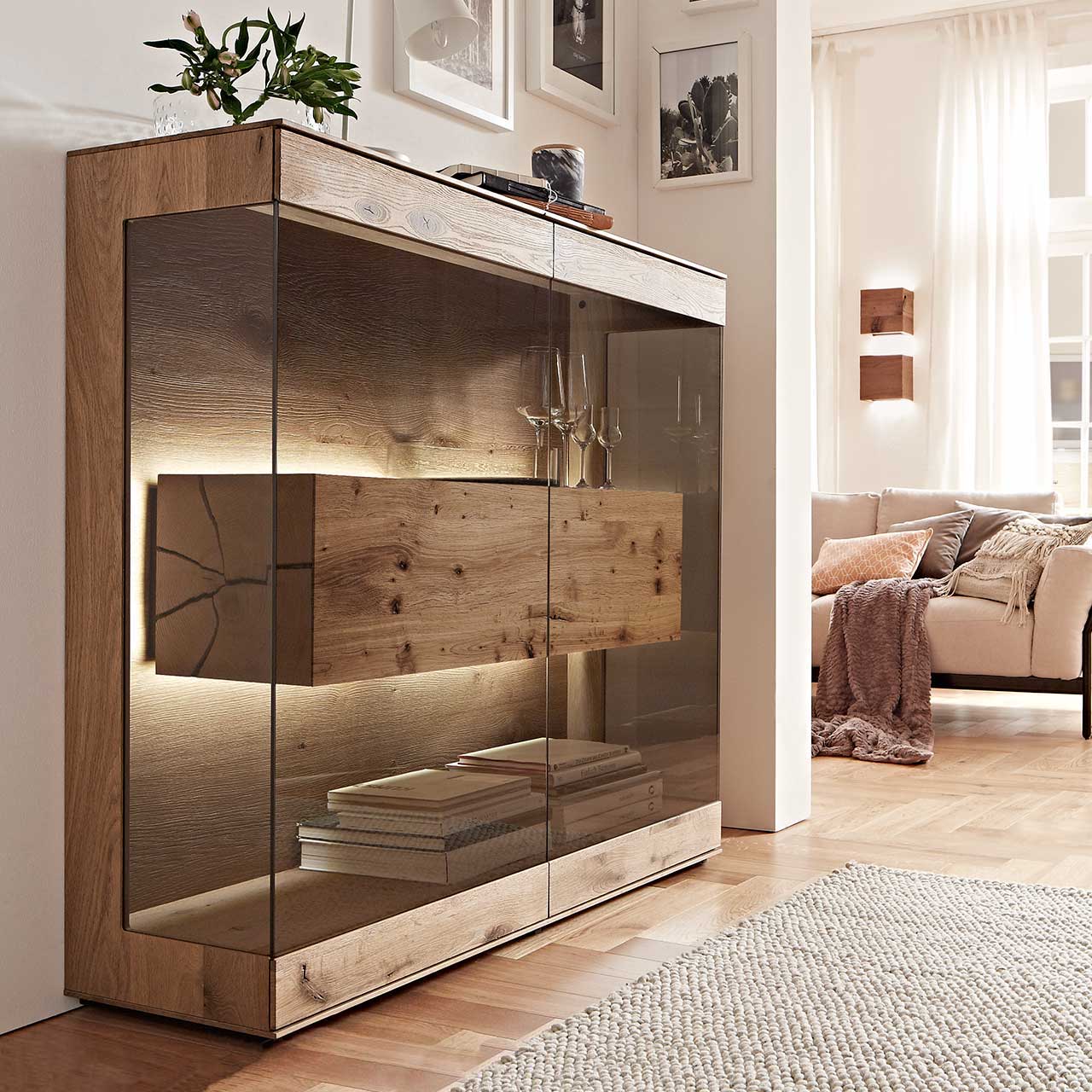 Milieubild Hartmann CAYA - Highboard mit Glasfront 7170-HM-6139 Edles Highboard aus massiver Kerneiche Umato mit großen über Ecke verlaufenden Glastüren in Parsolbronze. Innenliegend ein großer, beleuchteter Hirnholzblock mit Doppelschubkasten - in einem modernem Flur stehend.
