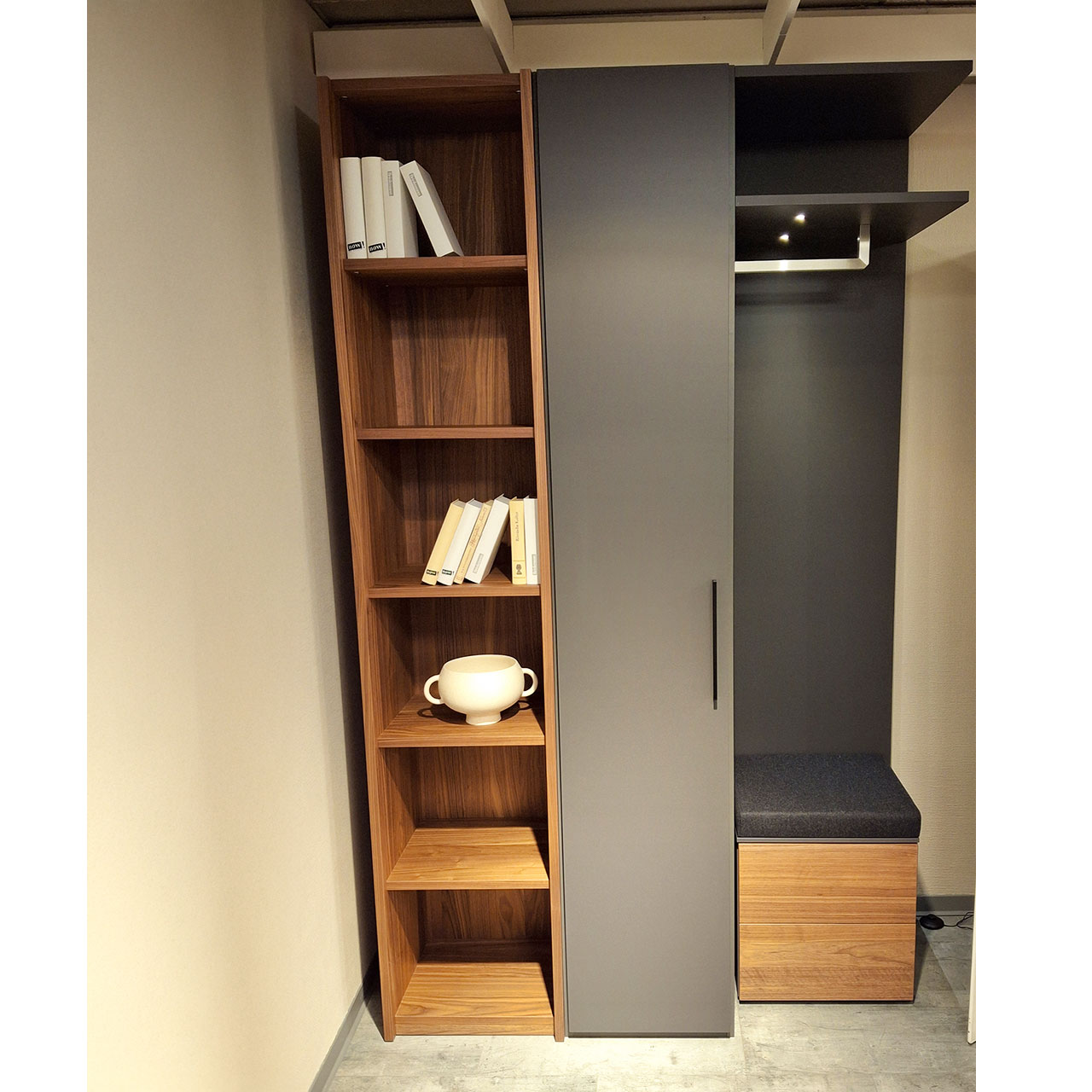 3-teiliger Garderobenschrank mit offenem Regal in Kernnussbaum links, mittig Regalschrank mit Tür in grau und rechts mit Anstellregal mit Beleuchtung und Sitztruhe unten.