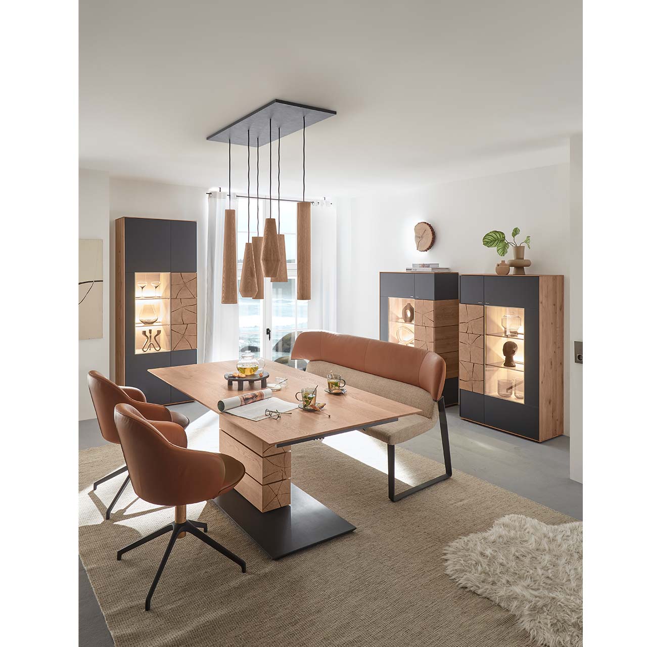 Milieubild - Hartmann CAYA - Standvitrine 7170-0087A li. Modernes Esszimmer mit rechteckigem Massivholz Tisch mit Sitzbank und zwei Stühlen mit Lederbezug in cognac. Über dem Tisch hängt ein Pendelleuchte mit Holz-Lampenschirmen. Im Hintergrund stehen zwei passende Highboards und eine Standvitrine in Anthrazit.