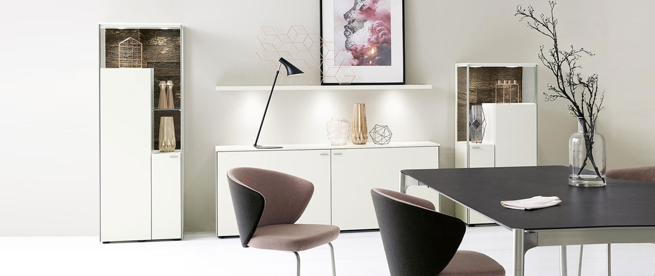 MH_banner_marken_venjako Ambientebild eines Wohn-Essbereiches der Marke Venjakob. Das Bild zeigt den Ausschnitt eines Esstisches mit zweifarbigen Stühlen. Im Hintergrund ein Highboard, ei nLowboard und eine Vitrine in Weiß mit Glasakzenten.