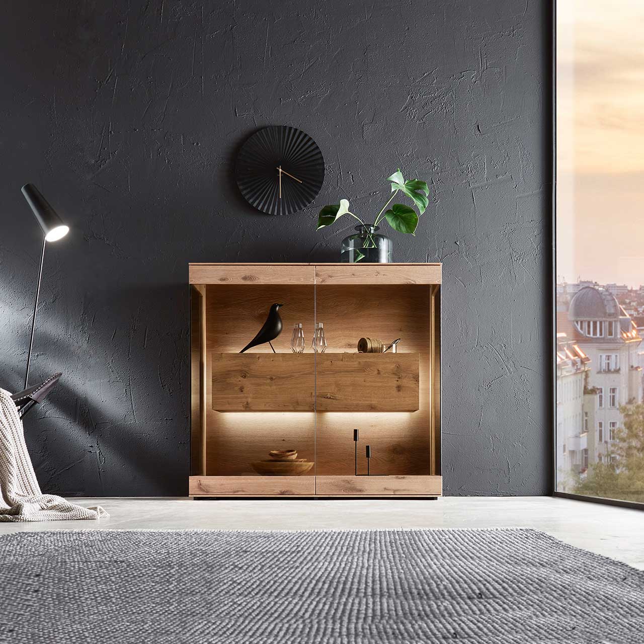 Milieubild Hartmann CAYA - Highboard mit Glasfront 7170-HM-6139 Edles Highboard aus massiver Kerneiche Umato mit großen über Ecke verlaufenden Glastüren in Parsolbronze. Innenliegend ein großer, beleuchteter Hirnholzblock mit Doppelschubkasten - an dunkelgrauer Wand stehend.