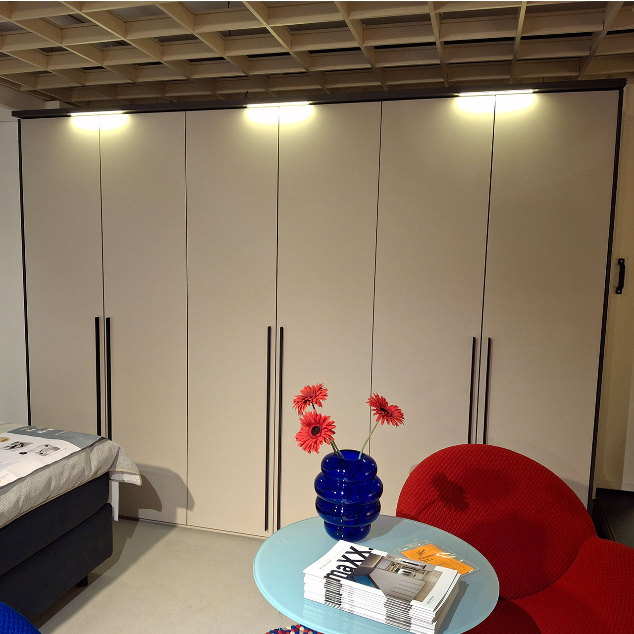 6-türiger Kleiderschrank mit LED-Beleuchtung über jeder Doppeltür. Rechts vor dem Schrank steht ein roter Sessel und ein Beistelltisch mit Glasplatte. In der vorderen linken Bildhälfte ist eine Ecke eines Bettes zu erkennen. 