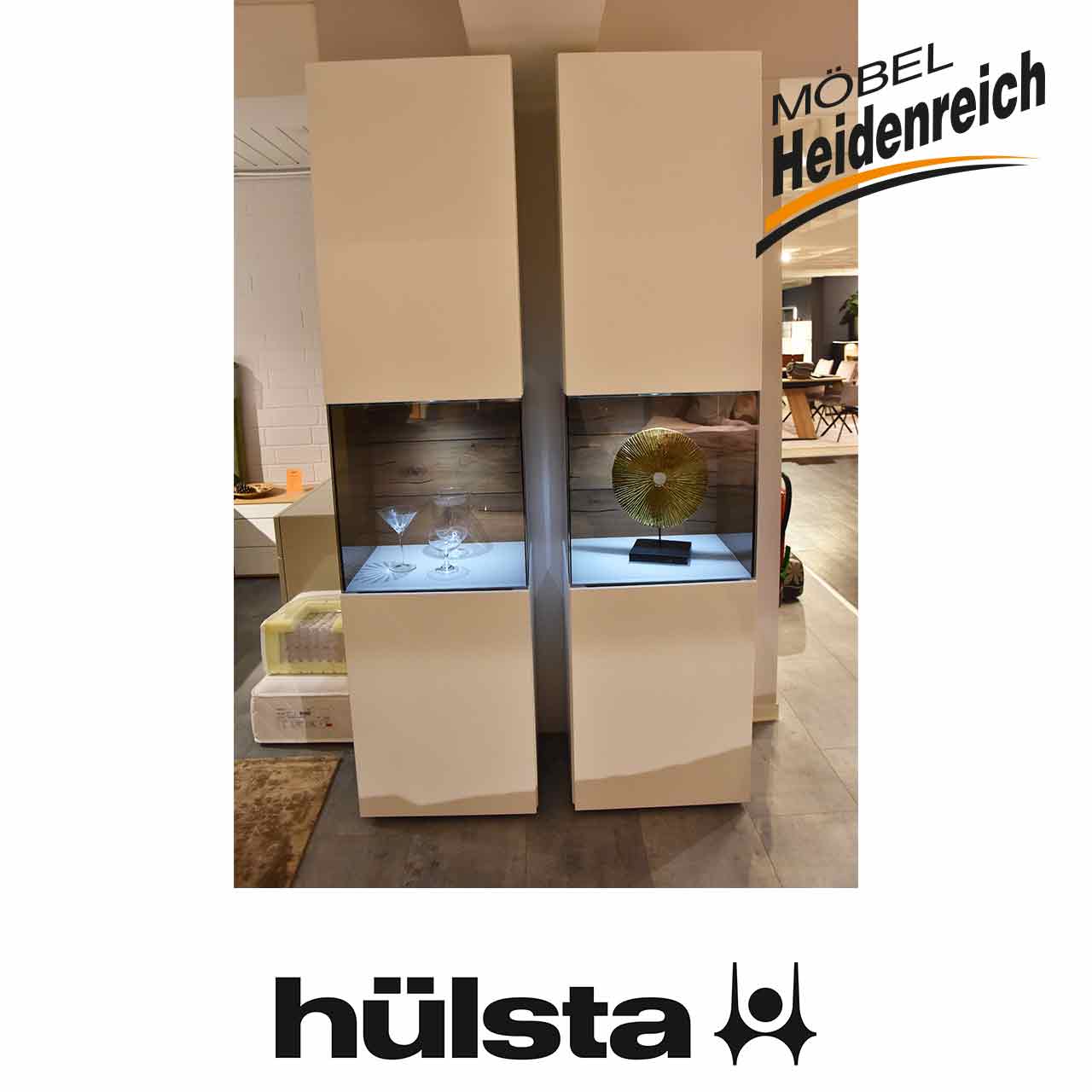 Zwei weiße Vitrinen nebeneinander. Hinter dem beleuchteten quadratischen Glasausschnitt in der Mitte befindet sich eine rustikale dunkle Holz-Rückwand.