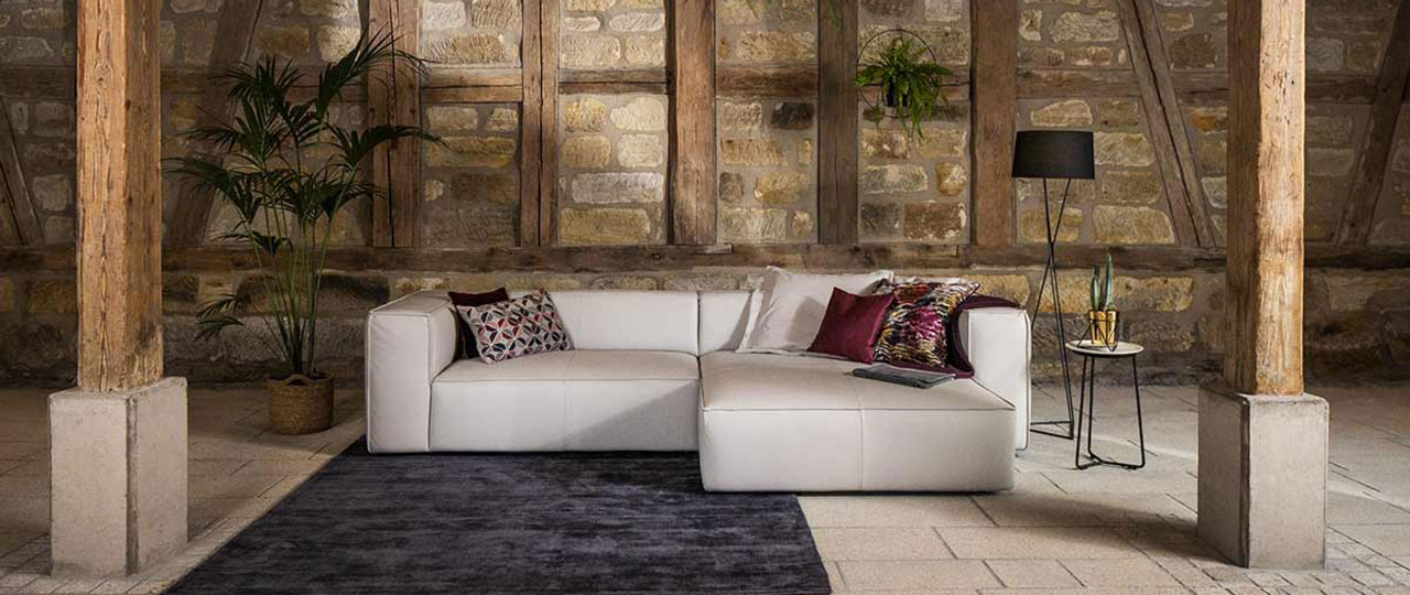 MH_banner_marken_schillig Ambientebild einer Sofalandschaft der Marke Schillig in Creme Leder. Das Sofa steht in einem luftartigen Raum mit offener Mauer / freigelegten Gebälk.