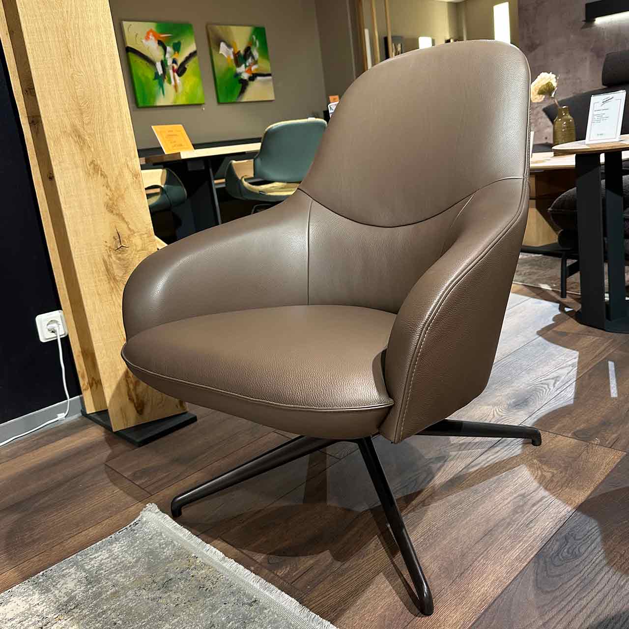 Der Sessel aus hellbraunem Leder hat Armlehnen und steht auf einem schwarzen sternförmigen Fuß.