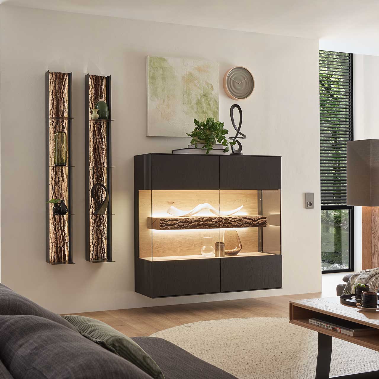 Milieubild Hartmann RUNA 8410 - Highboard 6122 und Wandregale 8024 - Kerneiche Natur Anthrazit gebeizt Modernes Wohnzimmer mit hängendem quadratischem Highboard aus Kerneiche Natur in anthrazit gebeizt, mit beleuchtetem Glasausschnitt und Rinden-Akzent. Links daneben zwei hängende Metallregale mit Rinden-Akzent.