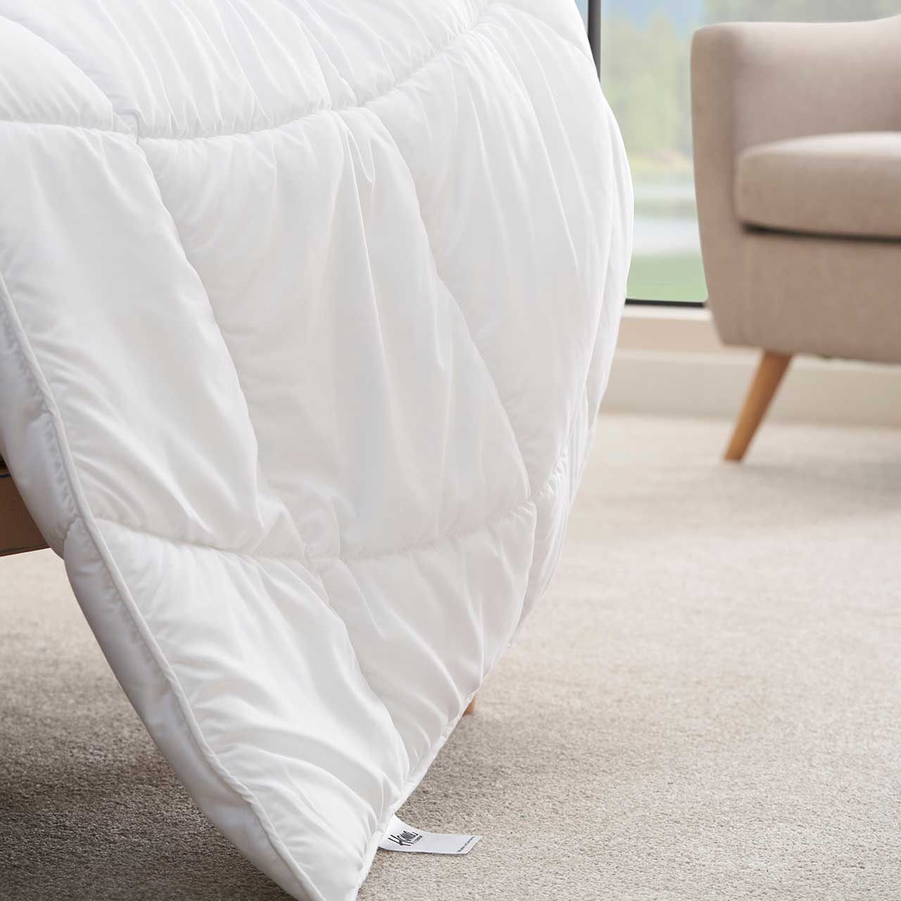 MH_Schlafzimmer_Bettdecken Produktbild Bettdecke, weiß, mit einem Teilausschnitt eines Sessels im Hintergrund.