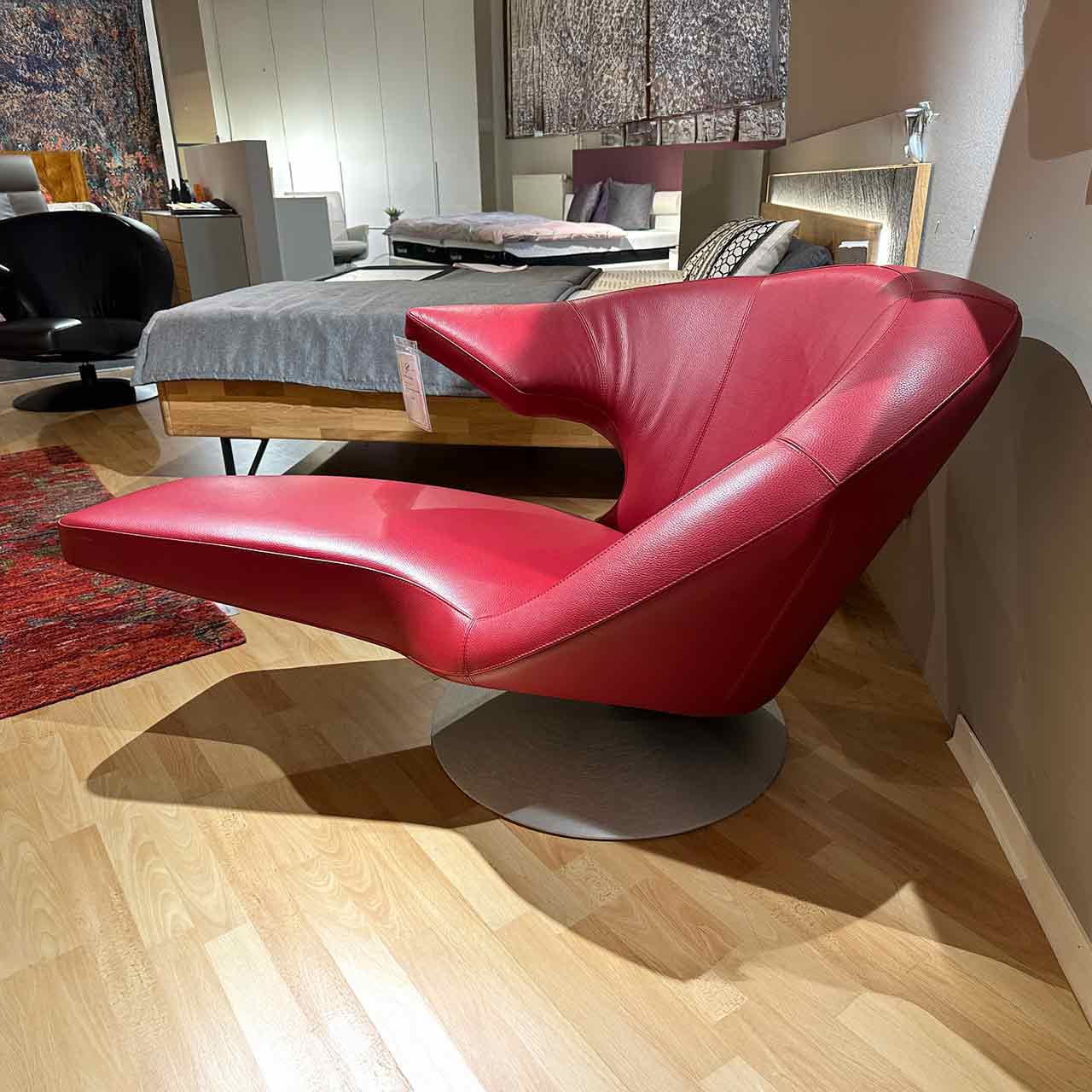 Drehsessel von der Seite: Auf der linken Seite steht eine prominente Armlehne hervor, das Fußteil ist liegenartig fixiert. Sessel steht auf rundem Teller.